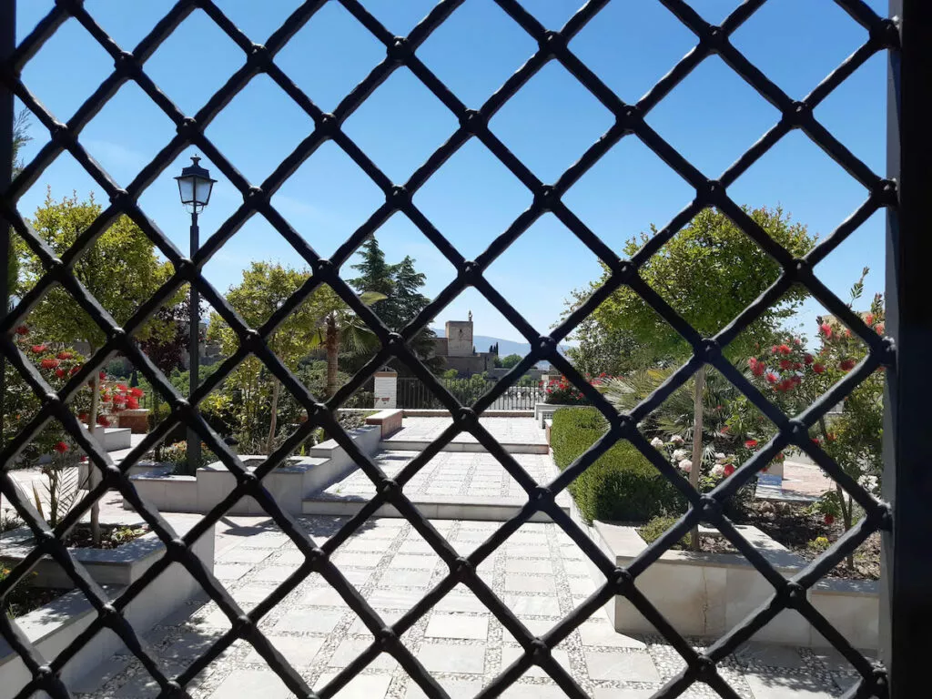 El jardín de la mezquita visto a través de una celosía de forja, con la torre de la Alhambra al fondo