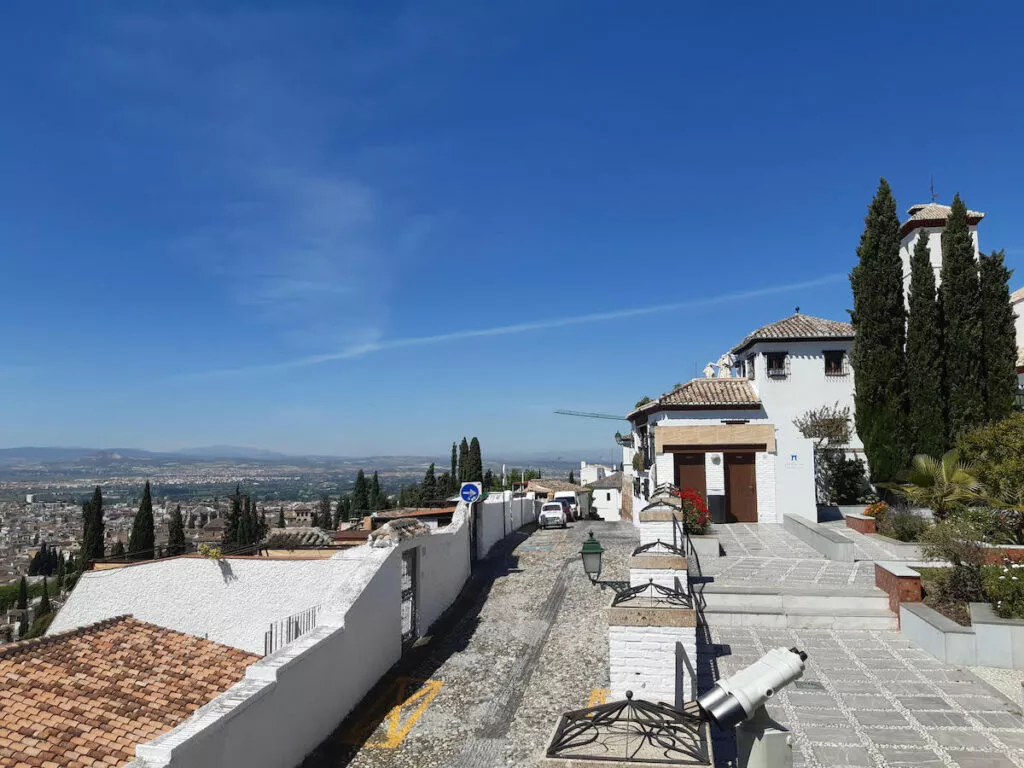 La calle empedrada junto a la mezquita, con la vega de Granada al fondo y un telescopio del mirador en primer término