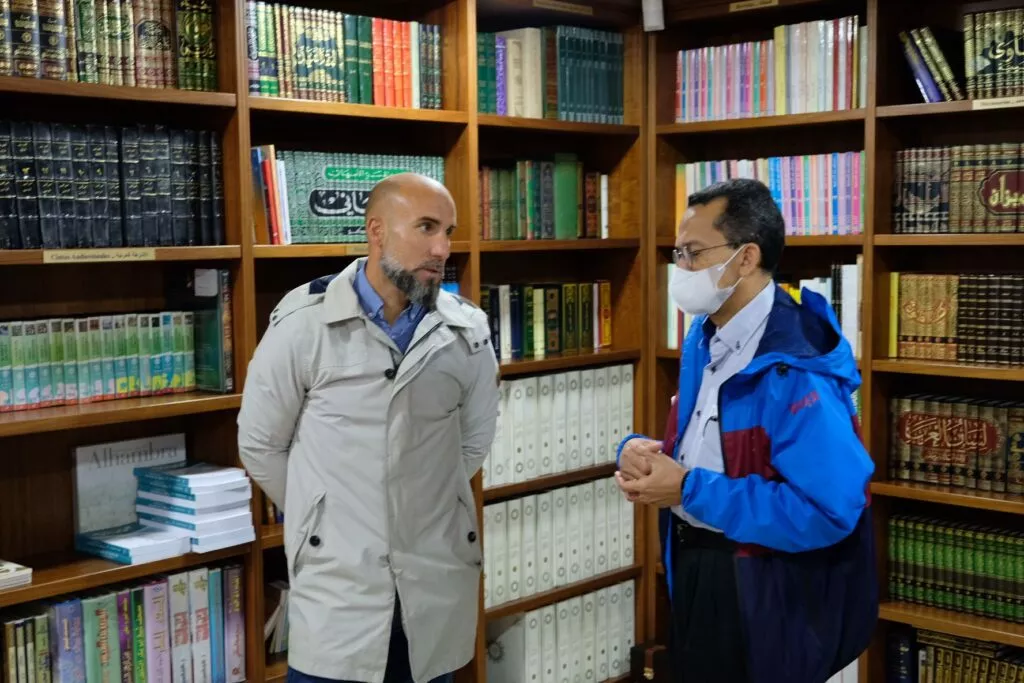 Un anfitrión conversa con un visitante ante las estanterías de la biblioteca de la mezquita