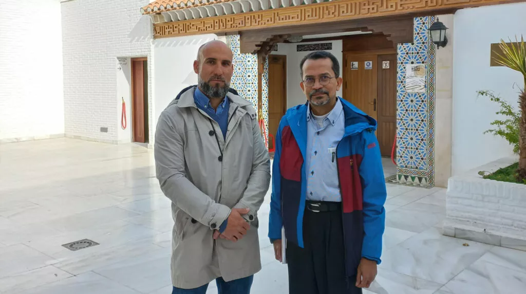 Dos hombres posan en el patio de mármol de la mezquita ante la puerta de madera y el alicatado
