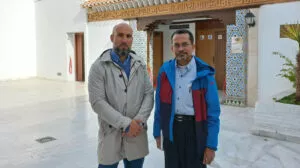 Dos hombres posan en el patio de mármol de la mezquita ante la puerta de madera y el alicatado