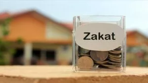 Bote de cristal lleno de monedas con una etiqueta que pone Zakat, con una casa desenfocada detrás