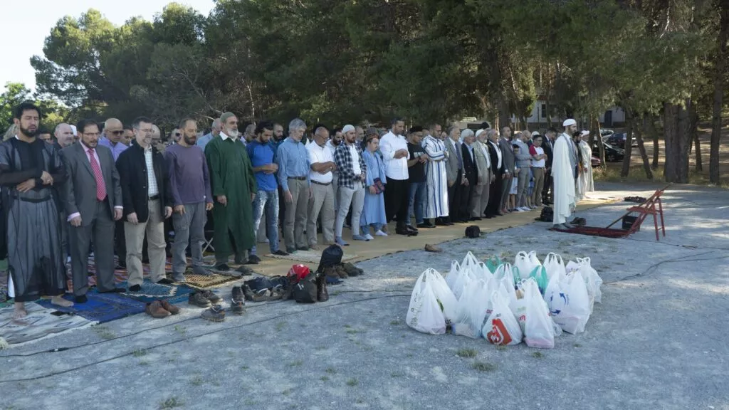 Oración del Eid al aire libre entre pinos, con los fieles en fila y bolsas de alimentos delante