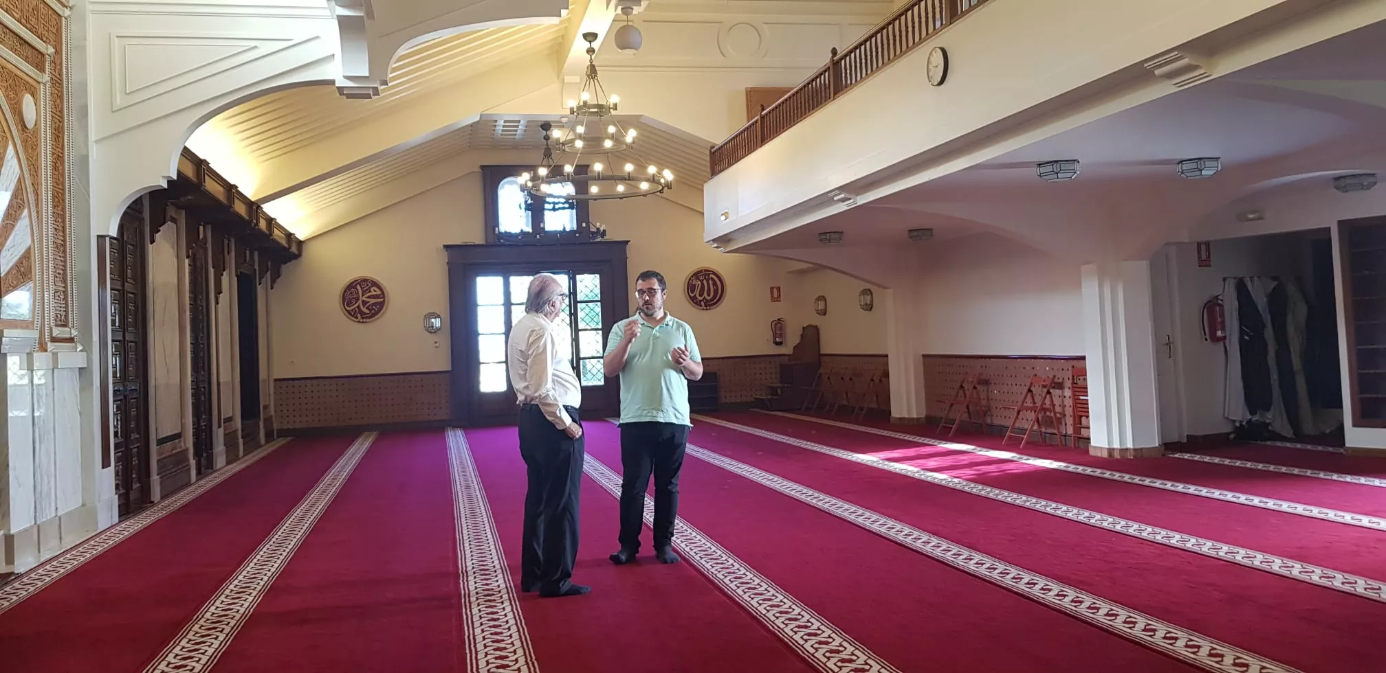 Dos hombres conversan de pie en la sala de oración vacía, sobre la alfombra roja