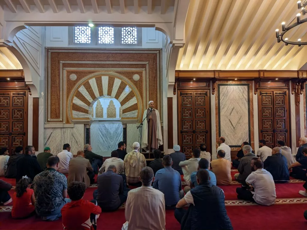 Sala de oración con alfombra roja, fieles sentados y el imam de pie ante el mihrab tallado