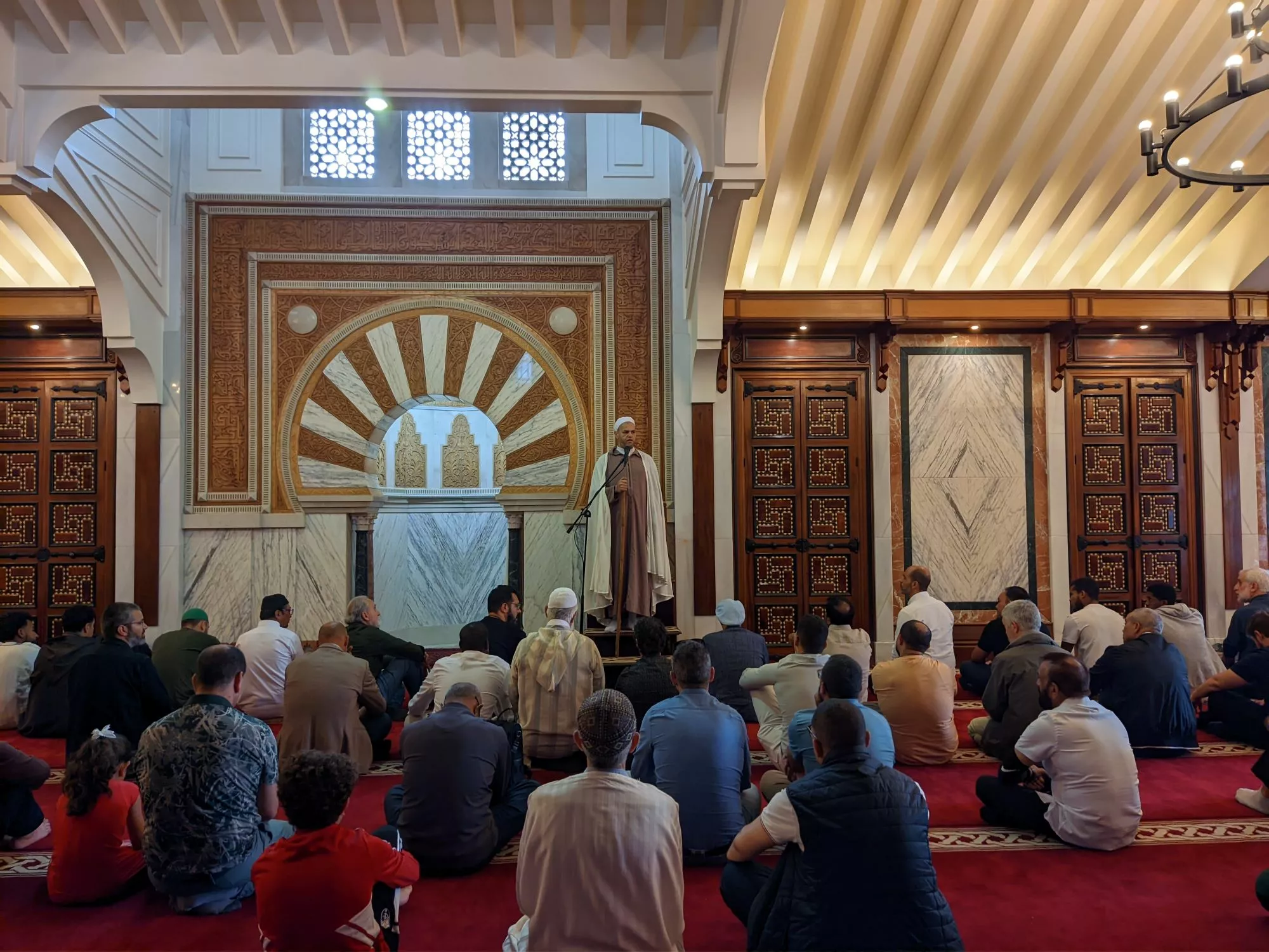 Sala de oración con alfombra roja, fieles sentados y el imam de pie ante el mihrab tallado
