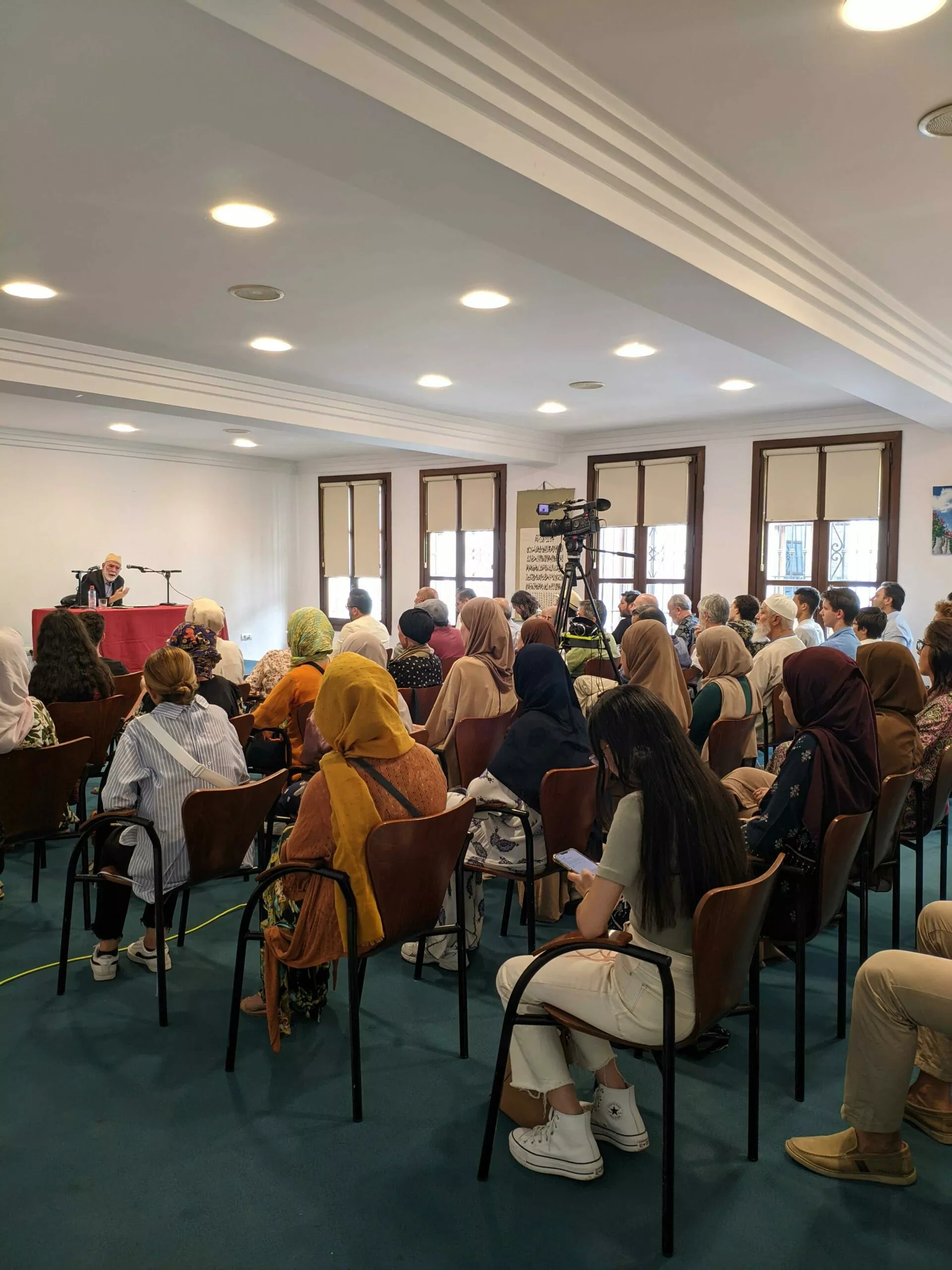 Sala llena de público de espaldas siguiendo la conferencia, con el ponente al fondo y una cámara grabando