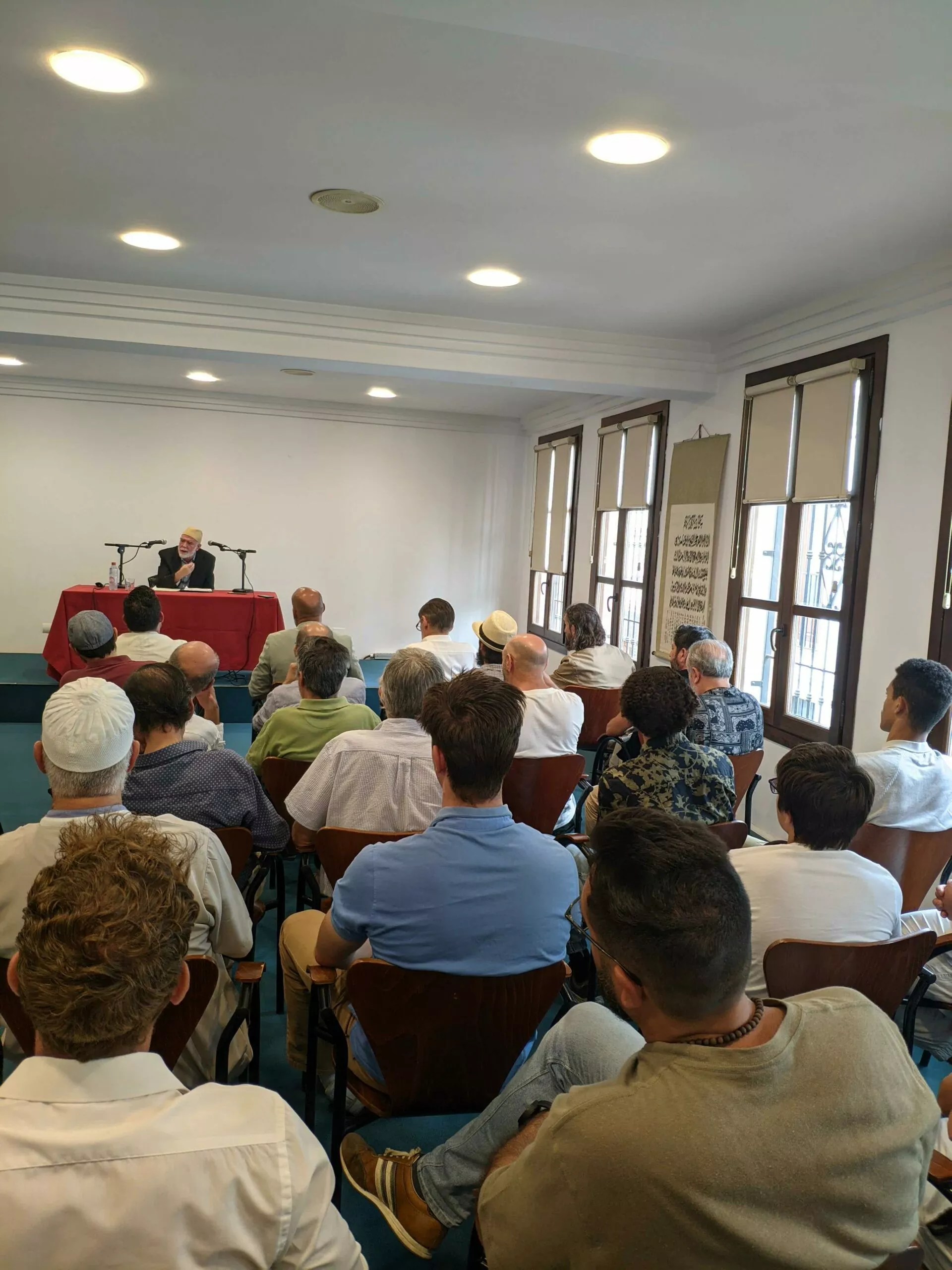 Público sentado de espaldas escuchando al conferenciante, que habla desde la mesa al fondo de la sala