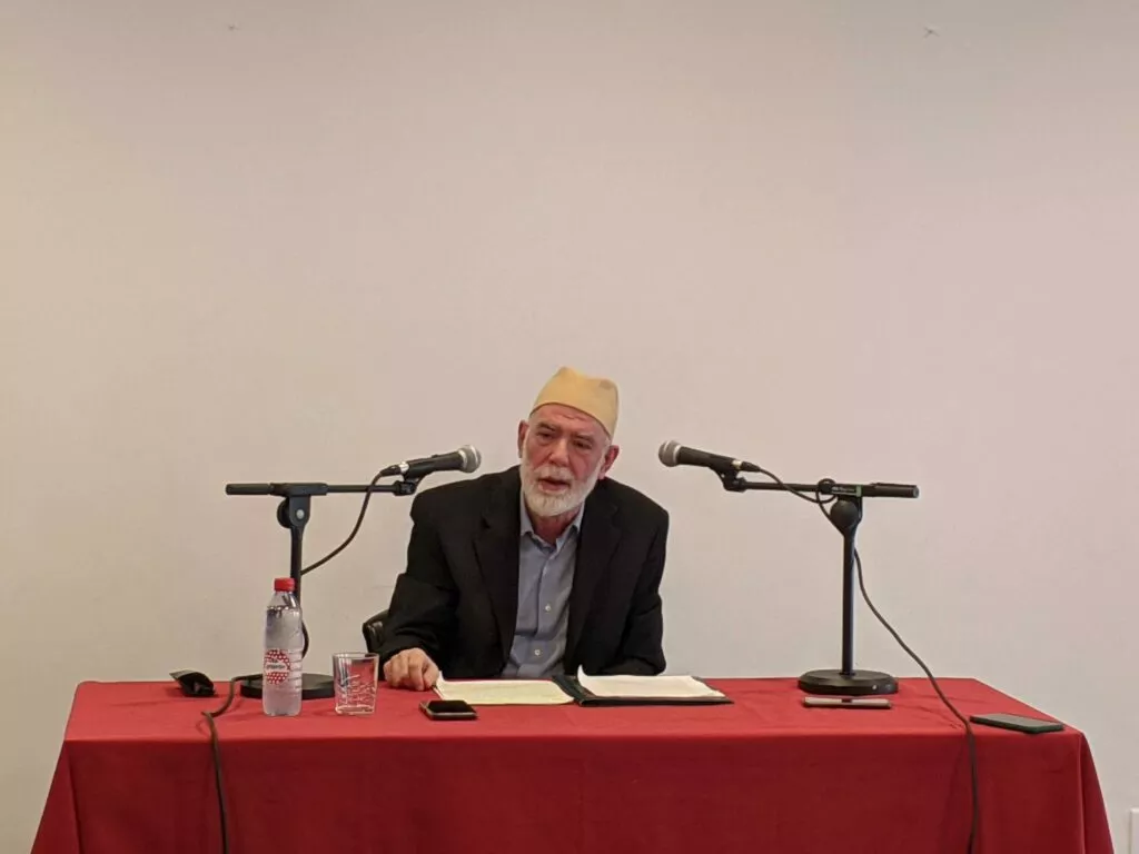 El conferenciante, con gorro amarillo y barba blanca, habla ante dos micrófonos en una mesa con mantel rojo