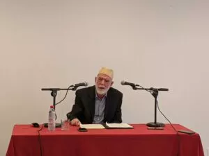 El conferenciante, con gorro amarillo y barba blanca, habla ante dos micrófonos en una mesa con mantel rojo