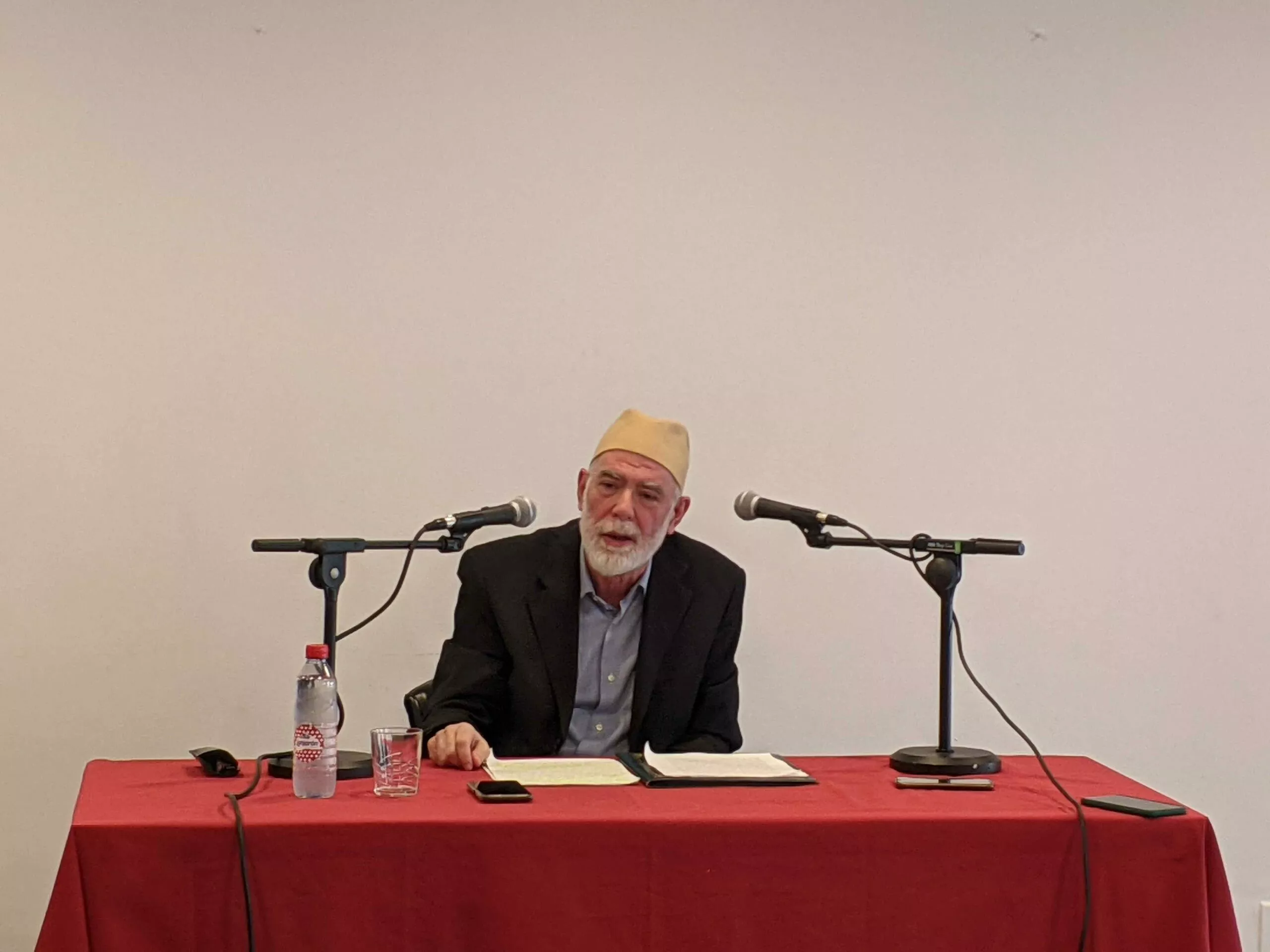 El conferenciante, con gorro amarillo y barba blanca, habla ante dos micrófonos en una mesa con mantel rojo