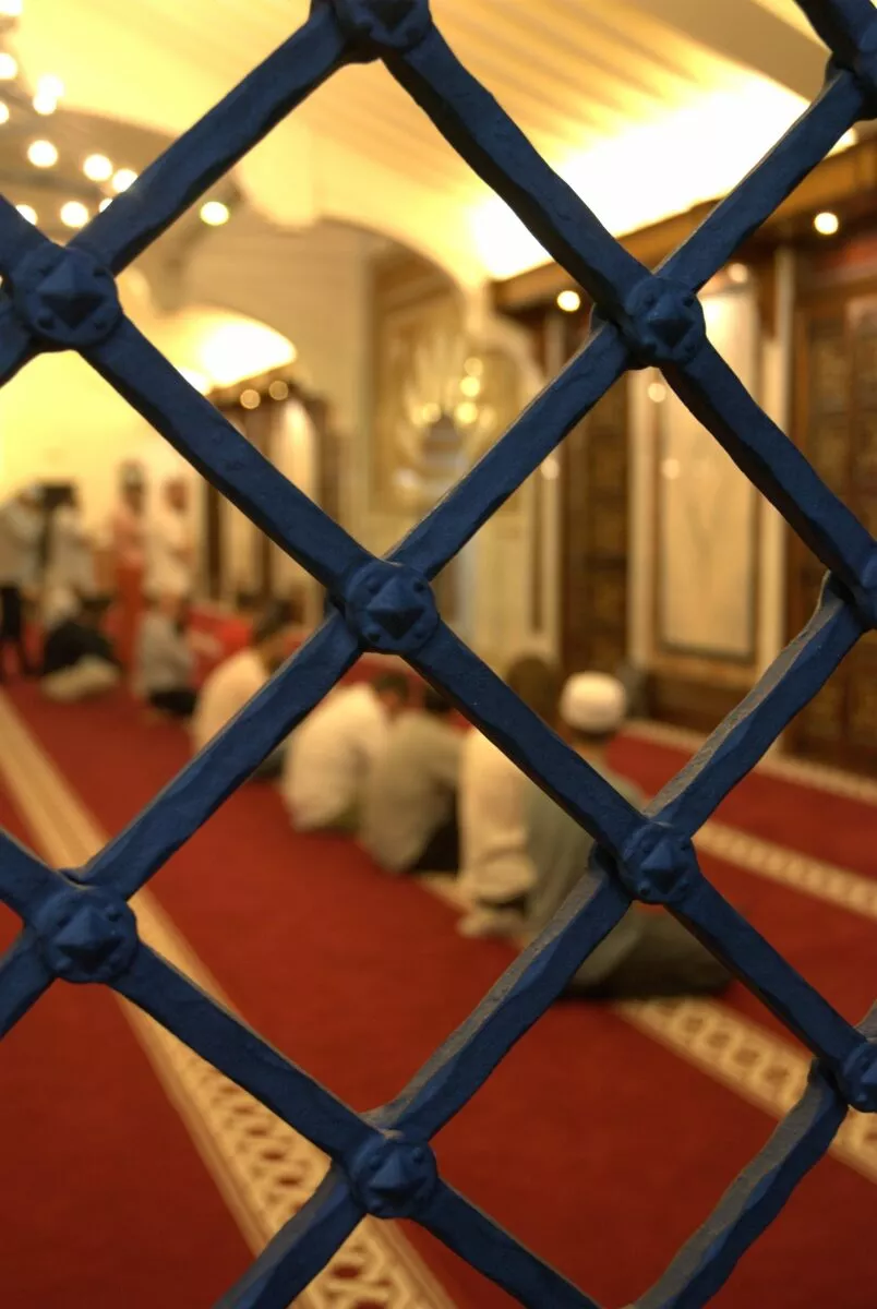 La sala de oración vista a través de una celosía de forja azul, con fieles sentados sobre la alfombra