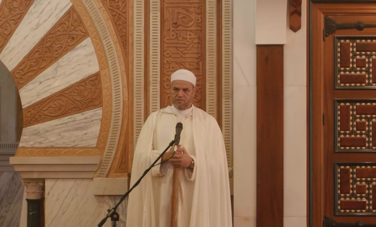 Fotograma del imam con turbante blanco y capa hablando ante el micrófono junto al mihrab