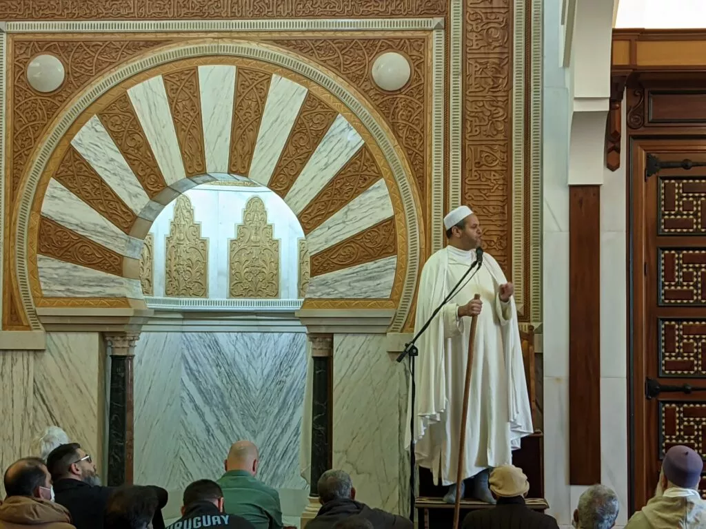 Imam con gorro blanco de pie en el minbar y fieles sentados ante el mihrab dorado