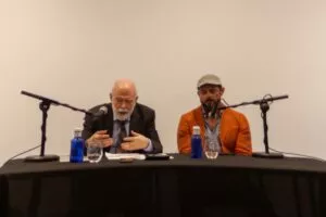 Ponente de barba blanca hablando ante el micrófono junto a otro con chaqueta naranja en la mesa