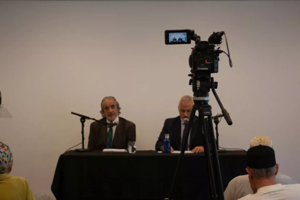 Cámara de vídeo sobre trípode filmando a los dos ponentes sentados a la mesa de la sala