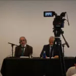 Cámara de vídeo sobre trípode filmando a los dos ponentes sentados a la mesa de la sala