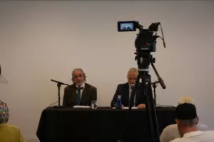 Cámara de vídeo sobre trípode filmando a los dos ponentes sentados a la mesa de la sala