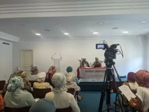 Dos ponentes en la mesa con el cartel del Mausim Granada ante un público de mujeres con pañuelos