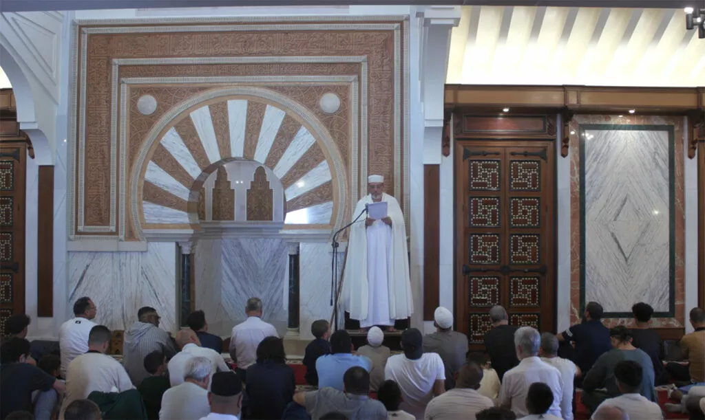 El imam, vestido de blanco, lee la jutba ante el mihrab con los fieles sentados en la sala