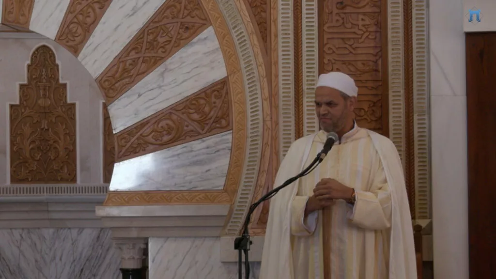 Predicador con capa blanca y chilaba de rayas hablando junto al mihrab de mármol