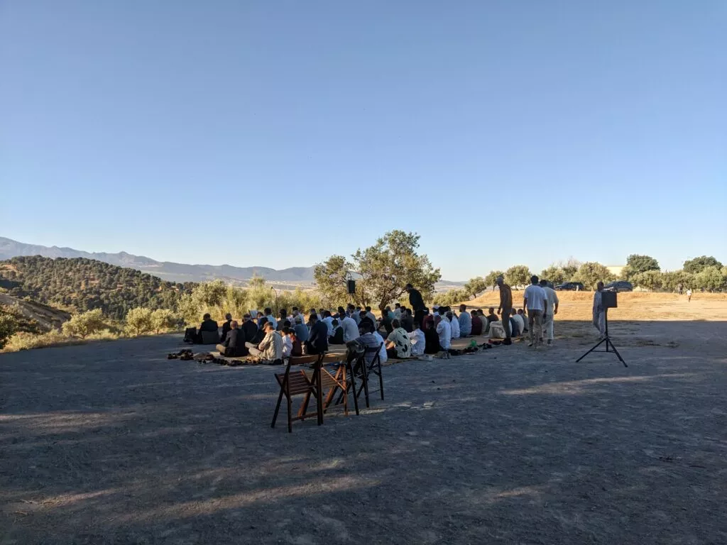 Fieles sentados en alfombras en una explanada al aire libre, con olivos y la sierra al fondo
