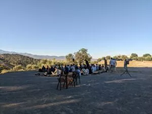 Fieles sentados en alfombras en una explanada al aire libre, con olivos y la sierra al fondo