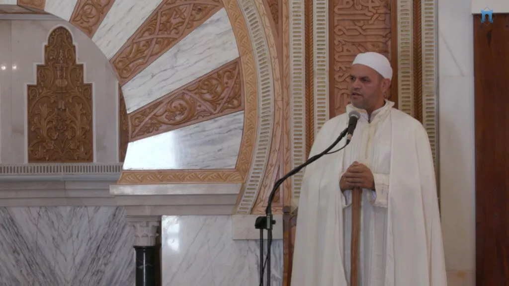 Imam de blanco hablando ante el micrófono, con el arco tallado del mihrab de fondo