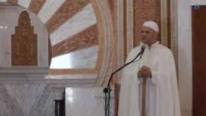 Imam de blanco hablando ante el micrófono, con el arco tallado del mihrab de fondo