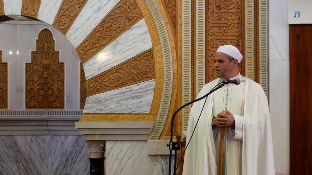 El imam con turbante y capa blancos sujeta su bastón mientras habla junto al mihrab