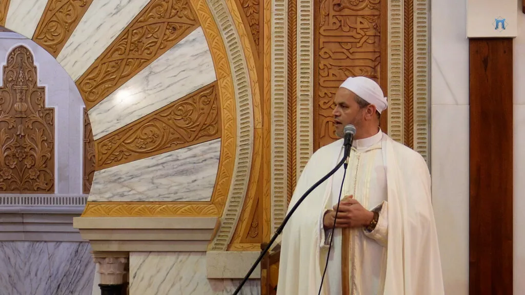 Primer plano del imam con turbante y capa blancos hablando ante el micrófono junto al mihrab