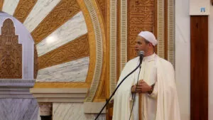 Primer plano del imam con turbante y capa blancos hablando ante el micrófono junto al mihrab