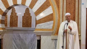 Imam con chilaba y turbante blancos dando la jutba junto al arco tallado del mimbar de mármol