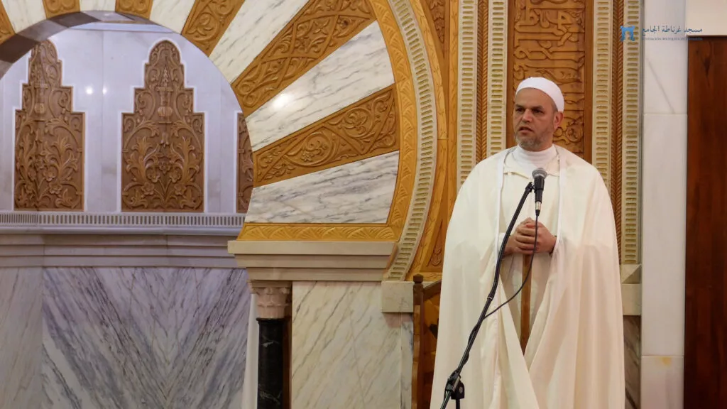 El predicador, de blanco, habla con las manos sobre el bastón junto al mihrab de mármol