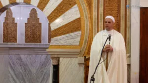 El predicador, de blanco, habla con las manos sobre el bastón junto al mihrab de mármol