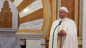 El imam, con turbante y capa blancos, apoyado en su bastón ante el arco dorado del mihrab