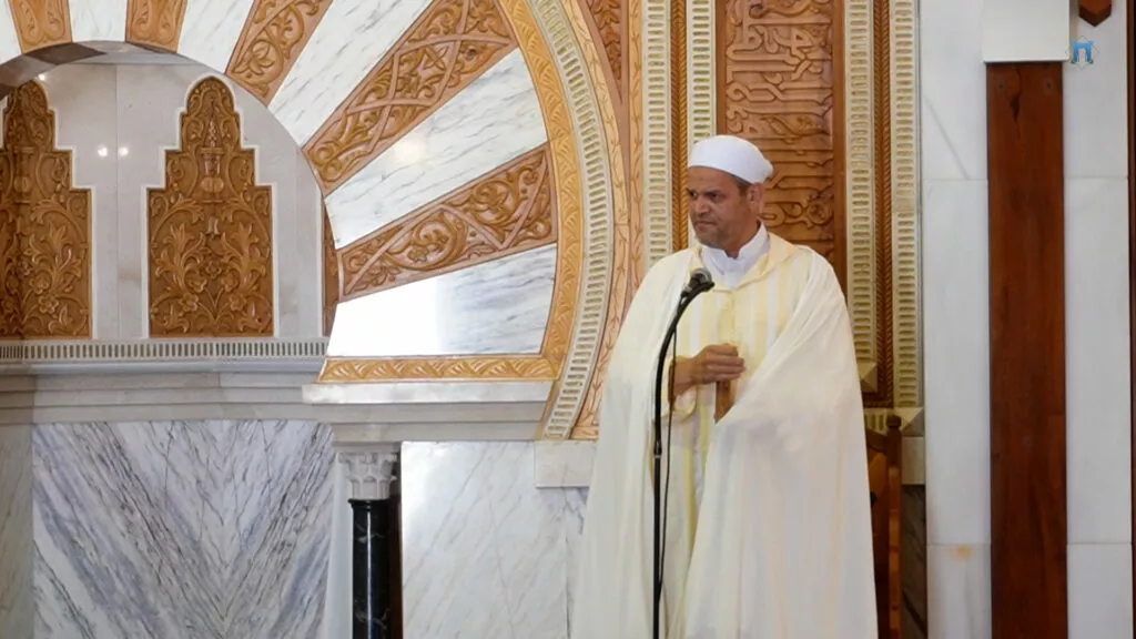 Imam con chilaba de rayas amarillas y capa blanca hablando ante el micrófono junto al mihrab