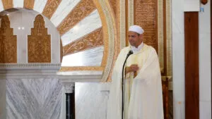 Imam con chilaba de rayas amarillas y capa blanca hablando ante el micrófono junto al mihrab