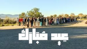 Filas de hombres rezando en una explanada entre olivos, con el rótulo Eid Adha Mubarak en árabe