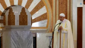Imam con turbante y capa blancos da la jutba apoyado en el bastón, junto al mimbar de mármol
