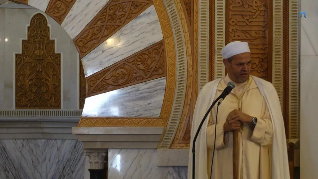 Imam con turbante blanco y chilaba clara predica ante el micrófono, junto al arco del mihrab