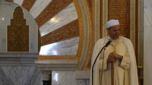 Imam con turbante blanco y chilaba clara predica ante el micrófono, junto al arco del mihrab