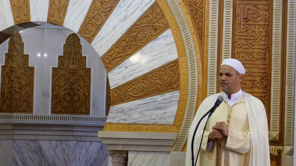 Jutba del viernes desde el mimbar, con el imam de chilaba clara y capa blanca ante el micrófono