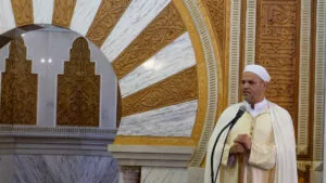 Jutba del viernes desde el mimbar, con el imam de chilaba clara y capa blanca ante el micrófono