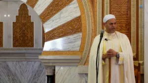 Imam con capa blanca y chilaba amarilla habla ante el micrófono junto al arco del mihrab