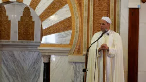 Predicador con turbante blanco, apoyado en el bastón del mimbar, habla ante el micrófono