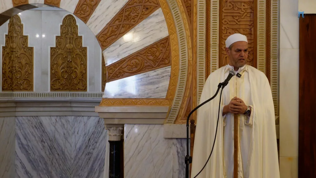 Jatib con chilaba y gorro blancos hablando ante el mihrab de mármol y yesería dorada