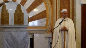 Jatib con chilaba y gorro blancos hablando ante el mihrab de mármol y yesería dorada