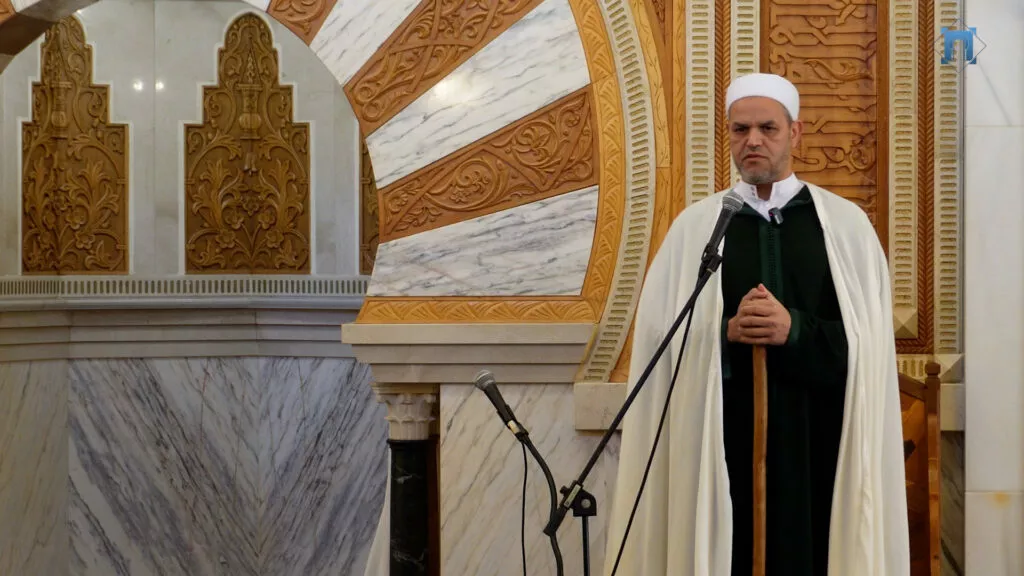 Imam con chaleco verde y capa blanca dando la jutba junto al arco tallado del mimbar