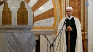 Imam con chaleco verde y capa blanca dando la jutba junto al arco tallado del mimbar